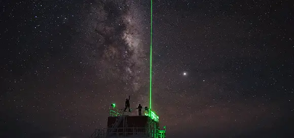 Long-exposure photograph of a bright green laser projecting from an observatory platform towards the brilliant core of the Milky Way.